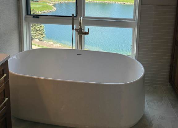 A white bathtub is in a bathroom with a window overlooking a large pond