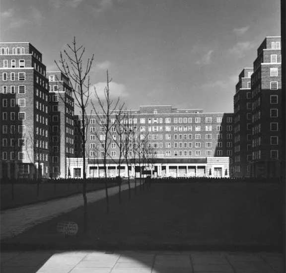 A black and white photo of a city street with buildings and a tree.