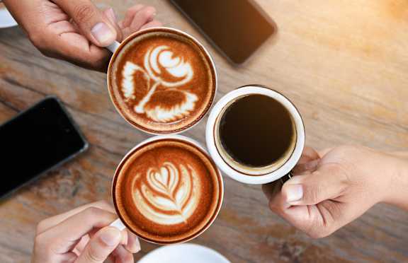 Three cups of coffee with latte art designs being held by people.