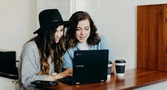 two women sitting at a table looking at a laptop