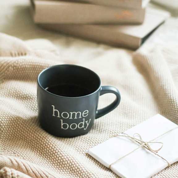 A black mug with the words 'home body' next to a stack of books.
