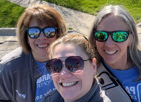 Three women wearing sunglasses and blue shirts are taking a selfie.