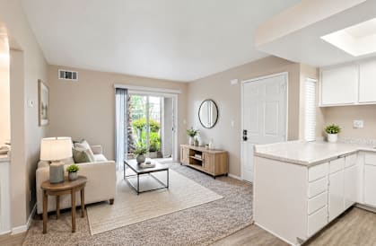 A well-lit, spacious living room with a white sofa, a wooden side table, and a large window.