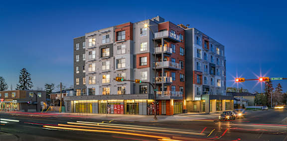A modern apartment building is lit up at dusk with cars passing by on the street.