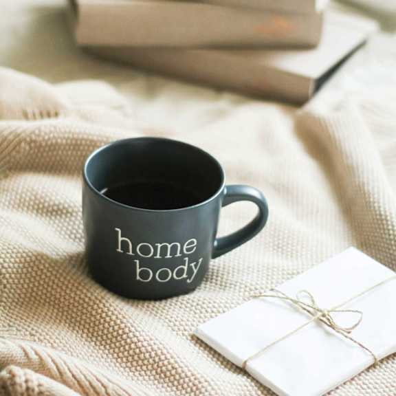 A green mug with "home body" written on it sits on a beige cloth next to a stack of books and a small white box tied with a string.