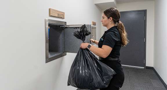 A woman is holding a black trash bag in front of a trash can.