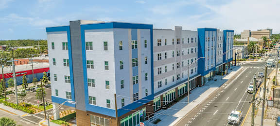 A modern multi-story building with blue and white exterior is situated on a street corner with cars parked along the curb.