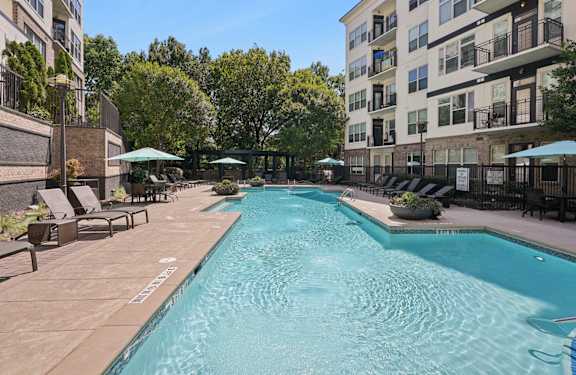 A pool surrounded by chairs and umbrellas in front of apartment buildings.