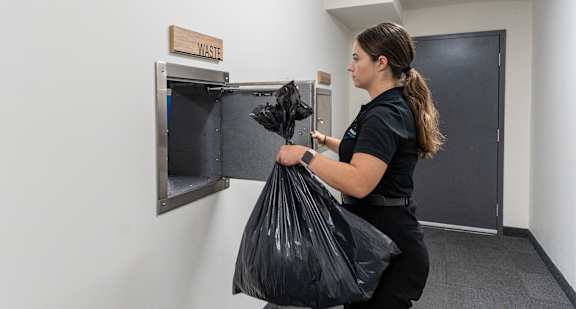 A woman is holding a black trash bag in front of a trash can.