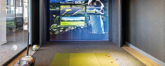 The golf simulator room at 3THIRTY3 Apartments features a large screen, green turf, and scattered golf balls for an immersive experience.