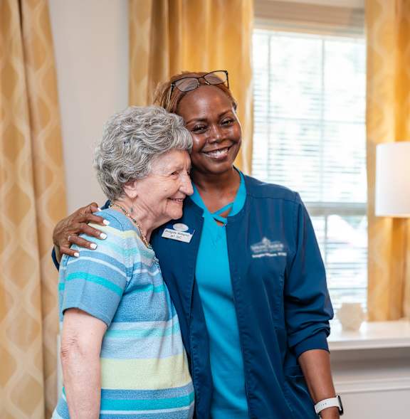 A nurse in blue scrubs is hugging an elderly woman in a striped shirt.