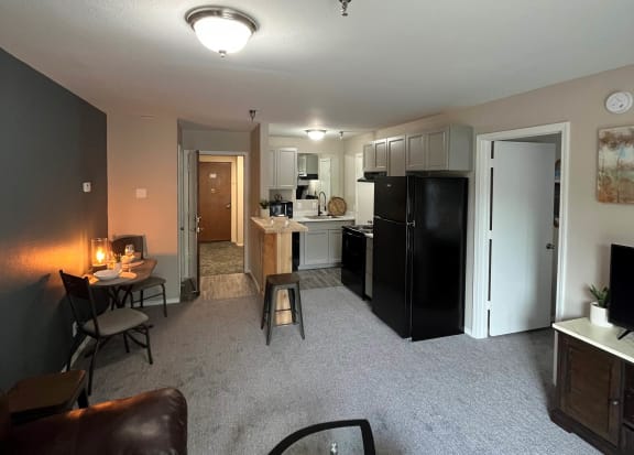 a living room with a couch a table and a chair and a kitchen with a refrigerator and at The Lodge Apartments, Tulsa, OK 74129