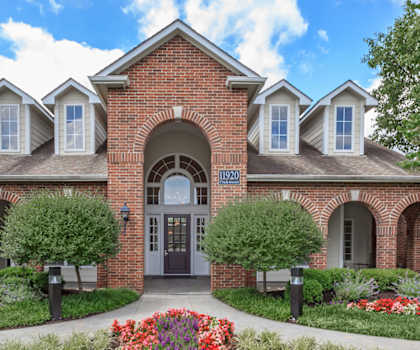 A red brick building with a black door and windows surrounded by greenery and flowers.