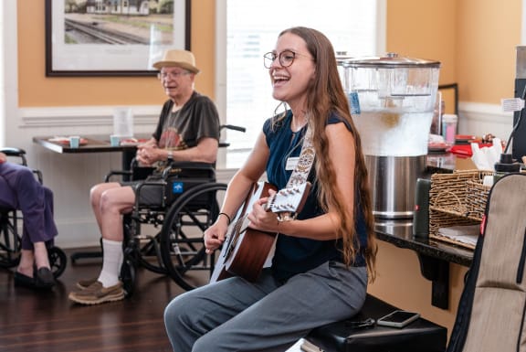 a woman sitting in a chair playing a guitar in a room with other people sitting