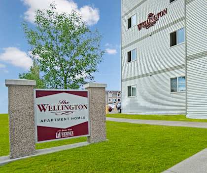 The exterior sign with trees and surrounded by greenery with the buildings in the background of Wellington Manor Apartment Homes. 