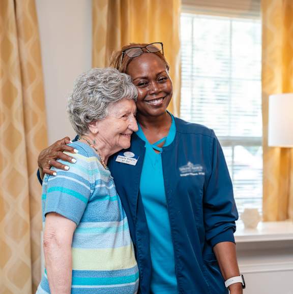 A nurse in blue scrubs is hugging an elderly woman in a striped shirt.