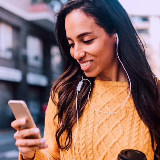 A woman in a yellow sweater is holding a phone and a coffee cup while wearing earphones.