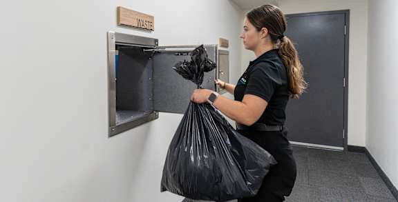 A woman is holding a black trash bag in front of a trash can.