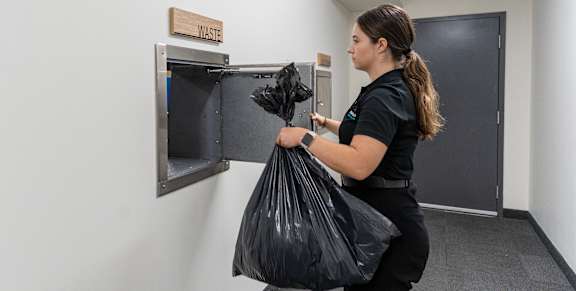 A woman is holding a black trash bag in front of a trash can. at G17 Apartments at Tamarack, Edmonton