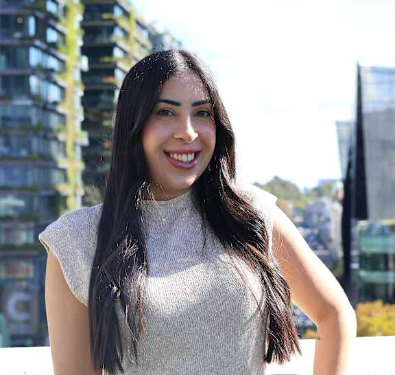A woman with long hair is smiling and wearing a white top and a brown belt.