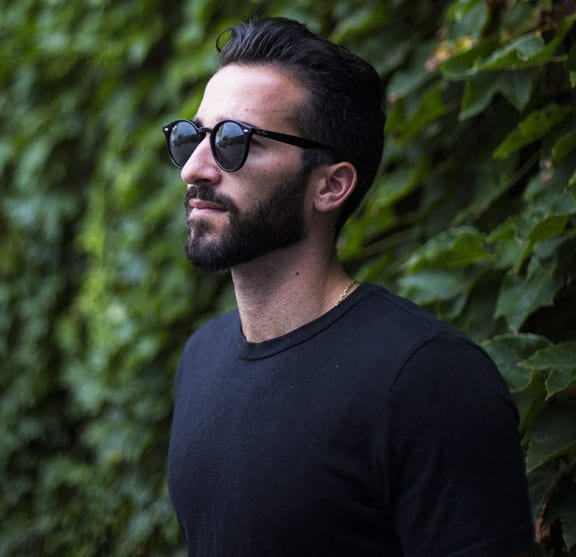 a man wearing sunglasses and a black tshirt standing in front of green plants