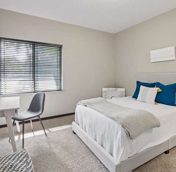 Bright staged apartment bedroom with carpet, upholstered bed, desk, window with blinds, and neutral finishes.