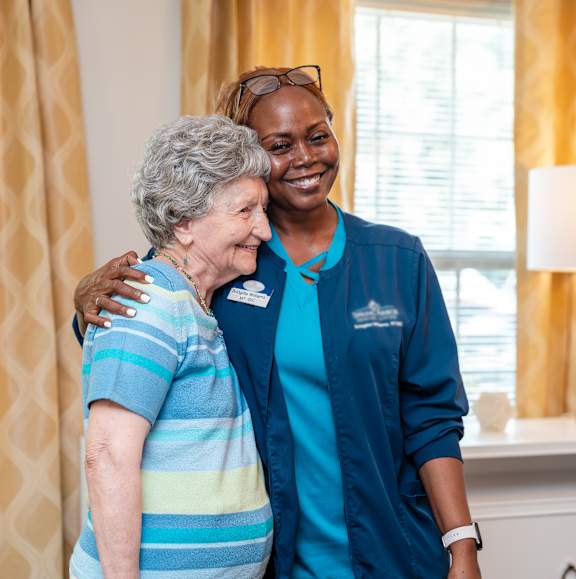A nurse in blue scrubs is hugging an elderly woman in a striped shirt.