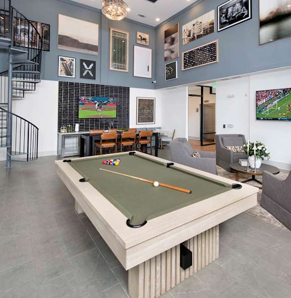 A billiard table is in the middle of a room with a grey couch and a TV.