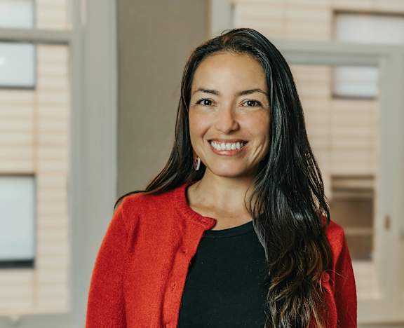 A woman with long dark hair wearing a red cardigan and black top standing in front of a window.