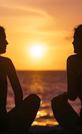 Two women sitting on the beach during sunset.