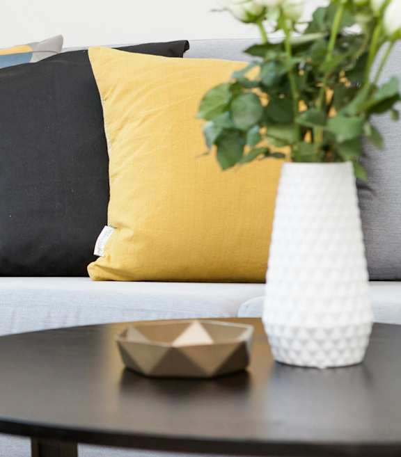 A grey couch with a yellow pillow and a vase with white flowers on a table.