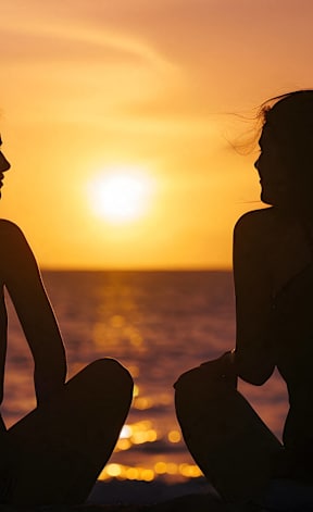 Two women sitting on the beach during sunset.