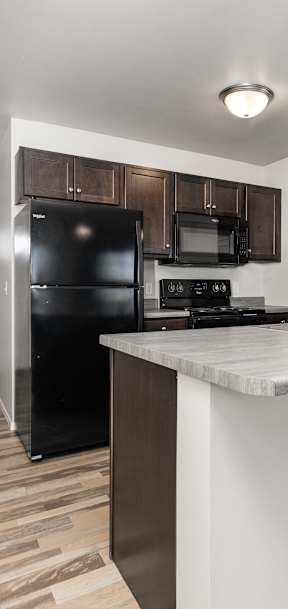 A kitchen with a black fridge and microwave, wooden floors, and a white countertop.