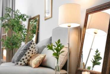 A living room with a grey couch, a table with a lamp, a vase with a plant, and a mirror.