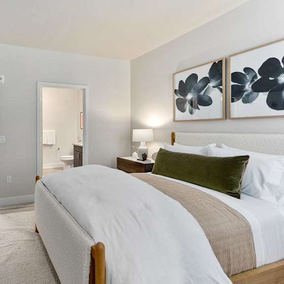A bedroom with a bed with white bedding, a rug, artwork, a bedside table, and a view of the bathroom at Dobson Ranch.