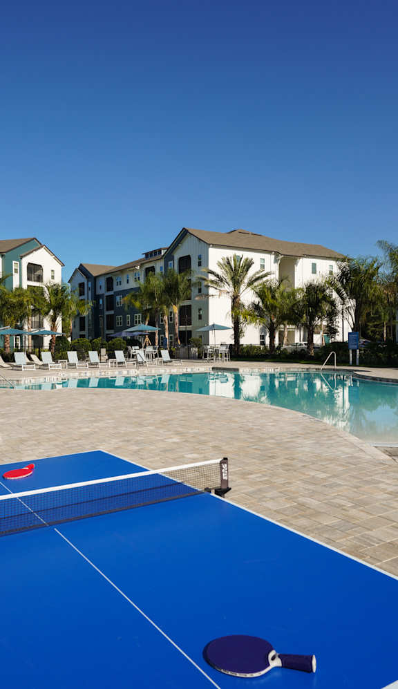 A blue table tennis court is surrounded by palm trees and buildings.