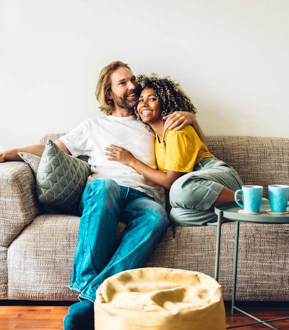 A couple is sitting on a couch with a potted plant to their left and a small table with cups on it to their right.