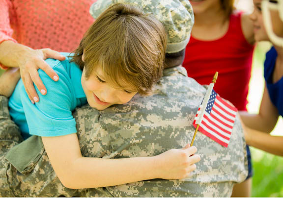 A child hugging a person in military uniform holding an American flag.