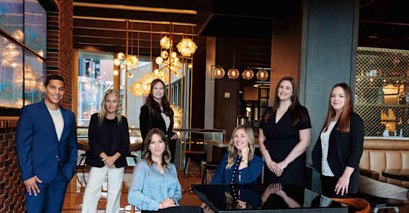 A group of people are standing and sitting around a table in a room with a brick wall and hanging lights.