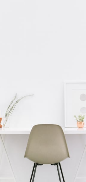 Tan chair in front of a white desk and white wall. The desk has a photograph and flowers on it.