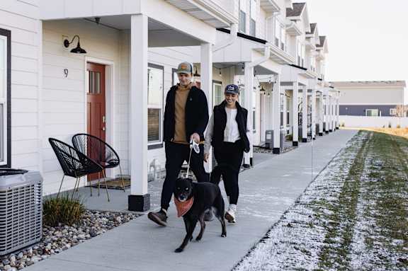 A man and a woman walking a dog down a snowy sidewalk.