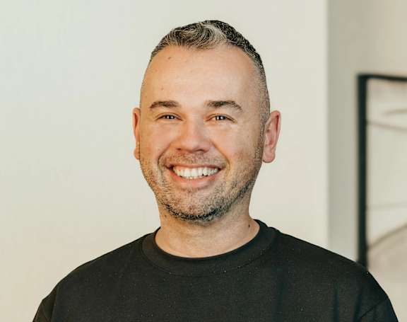 A man with a beard and short hair is wearing a black shirt and smiling.