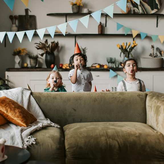 Two children wearing party hats are sitting on a couch in a living room.