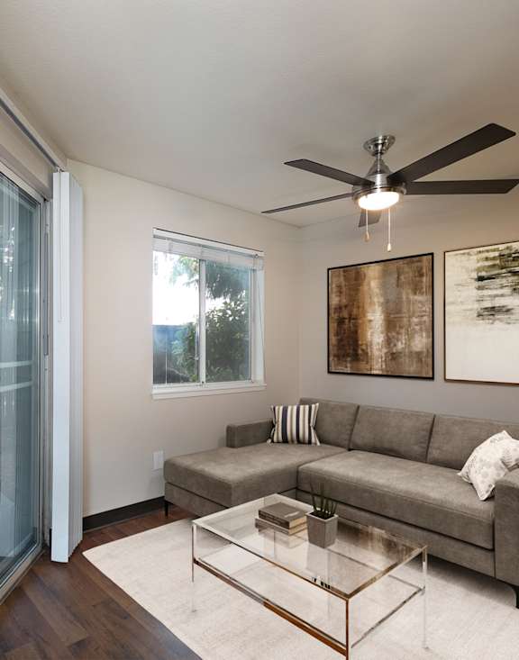 A modern living room with a grey sofa, a glass coffee table, and a ceiling fan.