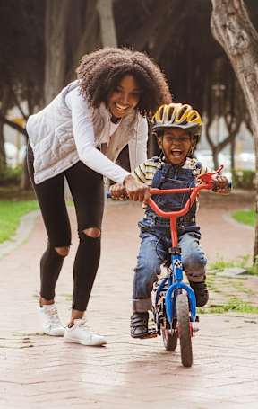 A woman helps a child ride a bike in a park.