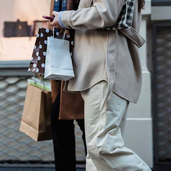 A person in a beige coat and white pants is holding shopping bags.