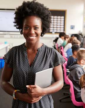 A woman is standing in front of a computer lab with students working on computers.