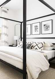 A white bed with a black frame is in a room with a white wall and a window with white curtains.