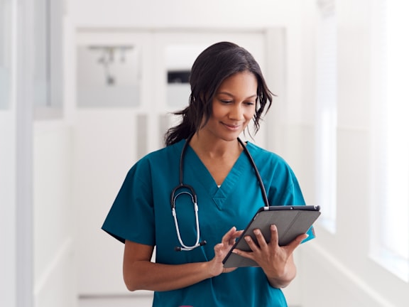 A woman in a teal scrub holding a tablet with a stethoscope around her neck.