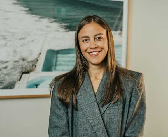 A woman in a grey suit stands in front of a framed picture of a boat.